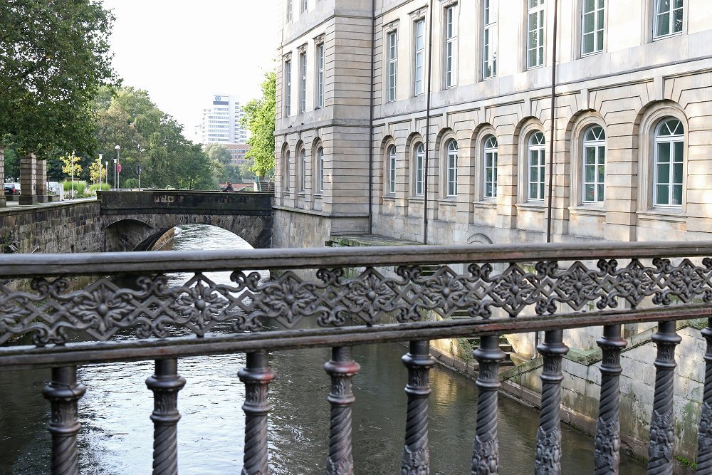 Zu sehen ist ein Fluss und eine daran angrenzende Häuserfassade. Fotografiert wurde von einer Brücke aus über ein metallenes, kunstvoll verziertes Geländer hinweg.